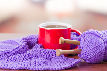 Obraz na płótnie Canvas Closeup purple yarn ball with knitting needles and scarf in progress lying on desk, coffee mug sitting next to it, blurry background