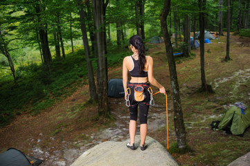 Rear view of young slim female climber standing on big natural boulder in the forest. Athletic girl is holding climbing rope and looking down on camping site. Climbing equipment.