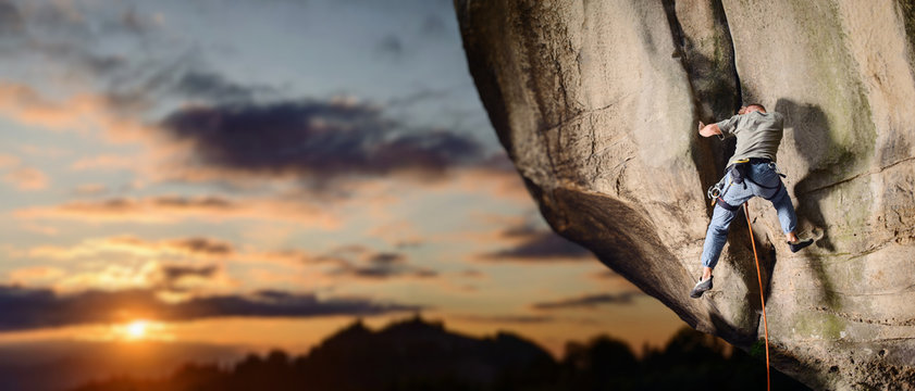 Young athletic male rock climber falling down from challenging route on overhanging cliff with rope and carbines on rocky wall against scenic sunset background. Summer time. Panoramic image