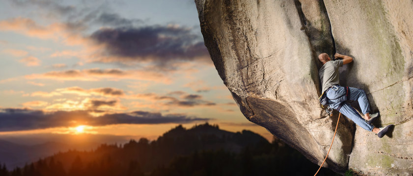 Young man rock climber climbing challenging route on rocky wall against scenic sunset background. Summer time. Climbing equipment. Panoramic picture