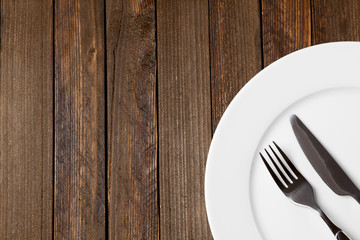 Fork and knife with white plate on dark wooden background, empty space on left