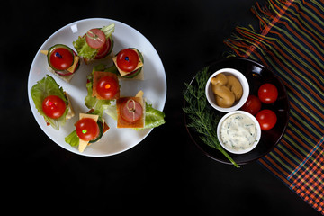 Small snacks canape with cherry tomatoes, cheeze, sausages and vegetables on bread on skewers on white plate with plate of sauces