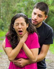 Young woman choking with man standing behind performing heimlich maneuver, park environment and casual clothes