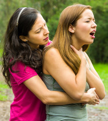 Young woman choking with lady standing behind performing heimlich maneuver, park environment and...