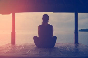 Silhouette of woman sitting and meditating near ocean at sunrise (intentional sun glare)