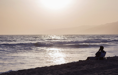 The silhouette of a man sitting on a sun lounger by the ocean. Light and shadow, a man rests during sunset on the beach.