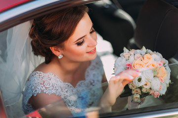 Gorgeous bride with beautiful smile is sitting in wedding car and holding summer flowers bouquet