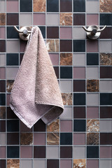 Small lilac terry towel for hands hanging on a towel rack in the bathroom on a square lined ceramic tile background.