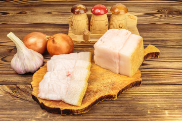 Piece and sliced fresh, raw pork lard on wooden board, garlic, onion on the table.