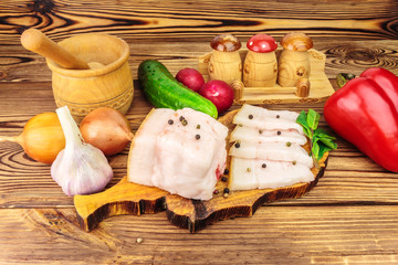 Piece and sliced fresh, raw pork lard on wooden board with vegetables, spices on the table.