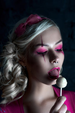 Beautiful blonde girl with two pigtails, with creative doll make-up: pink glossy lips, wearing pink skeleton dress licking lollipop. for the Halloween party. Close up, retouched image.