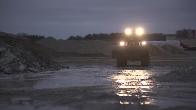 Excavator or bulldozer going along the construction site with ground  heaps or piles in the dark with headlights on. Slow motion wide shot.