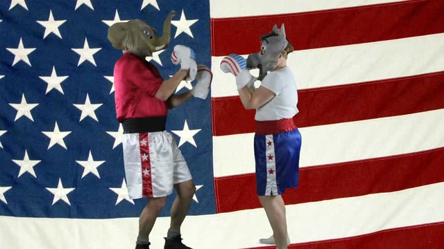 Man in elephant GOP mask and woman in donkey Democrat mask wearing boxing shorts sparring against against an American Flag .
