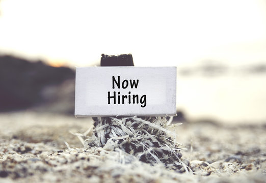 concept image, word NOW HIRING on white canvas frame with blurred beach and clam shell background.