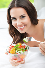 smiling young woman eating salad, on bed