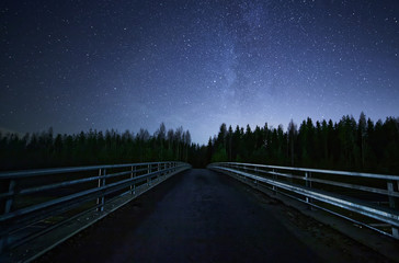 A road leading into night sky full of stars and visible milky way. A Bridge and dark forest on the foreground.