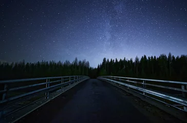 Deurstickers A road leading into night sky full of stars and visible milky way. A Bridge and dark forest on the foreground. © Teemu Tretjakov