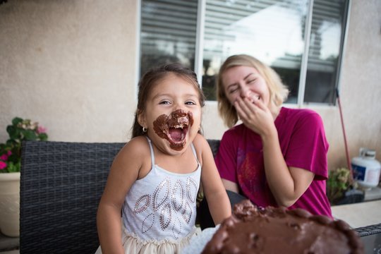 Young girl, chocolate cake on her face, sitting with family member, laughing