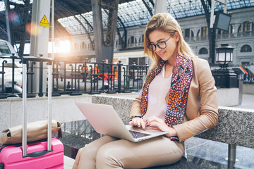 Fototapeta premium Beautiful Caucasian woman using net-book while sitting in train station interior. modern businesswoman using a laptop computer while waiting for the train. Flare light.