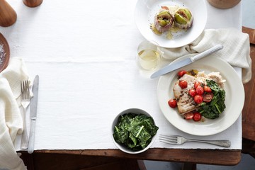 Overhead view of meal table with plate of prosciutto wrapped figs, wilted spinach, cherry tomatoes, tuna steak and capers