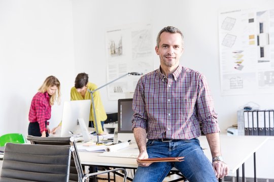 Mature man in office sitting on desk holding digital tablet looking at camera smiling