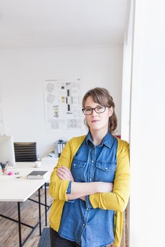 Young woman leaning against wall in office arms crossed looking at camera