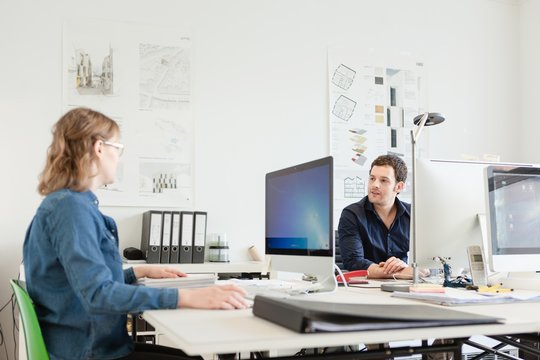 Colleagues sitting at desk in office using computers talking