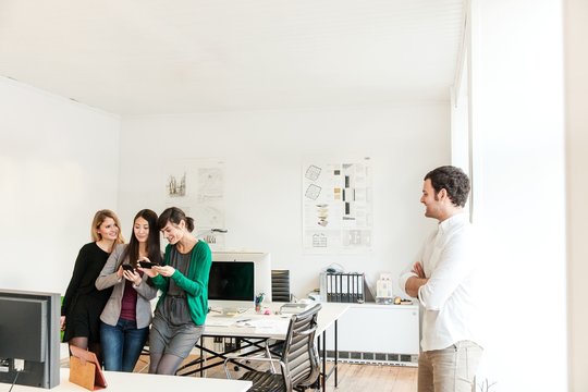 Mid adult man in office looking at colleagues using smartphones smiling