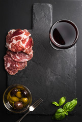 Glass of red wine, meat appetizer, olives and basil on black  slate stone board over dark background