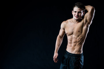 Fototapeta premium Attractive muscular young man looking to the camera and smiling with arm up, hand behind head, isolated on black background