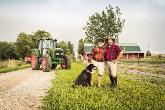 Father and son with dog on farm in front of tractor looking at camera smiling