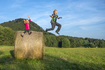 A young boy jumps off a bale of hay with her sister at summertime