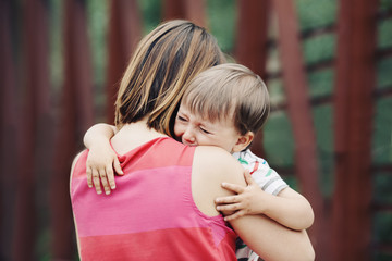 Portrait of  young Caucasian woman mother comforting her crying little toddler boy son outside in park on summer day, parenthood lifestyle concept