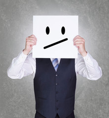 Geschäftsmann hält sich Schild mit genervtem Smiley vor sein Gesicht