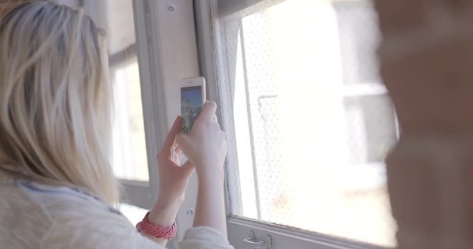 Over the shoulder view of beautiful young blonde woman using a smart phone to take pictures through a window in an urban hallway with brick walls. Hand held, slow motion, recorded in 4K at 60fps.