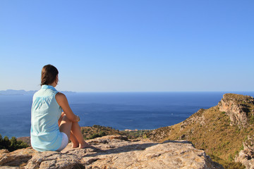 Fototapeta na wymiar Contemplative beautiful girl sitting on the rocks in front of the mediterranean sea meditating. Blue and orange tones on the picture. This photo transmit calm, meditation, relax, holidays.