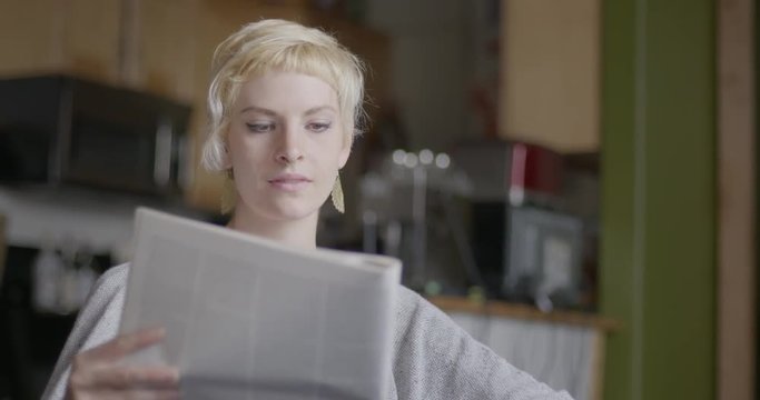 Close up of attractive young woman with short blonde hair reading a newspaper then looks up to camera and smiles. Urban loft location.  Hand-held camera recorded at 60fps.
