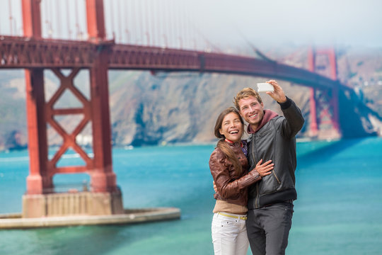 Happy young couple tourists taking selfie in San Francisco by Golden Gate Bridge. Interracial young modern couple using smart phone. Asian woman, Caucasian man.