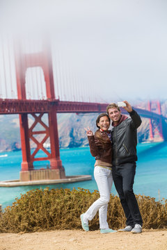 Tourists couple taking selfie photo in San Francisco by Golden Gate Bridge. Interracial young modern couple using smart phone by famous american landmark. Asian woman, Caucasian man.