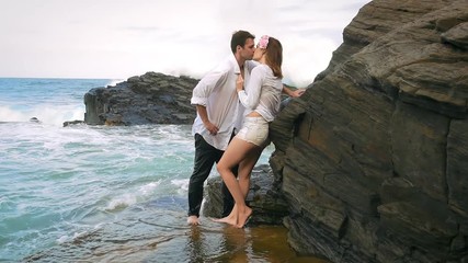 Young couple leaning against rocks kissing with waves crashing behind them