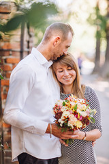 Close up fashion romantic portrait of young pretty couple kissing and hugs on the street at evening bright sunlight. Blonde gril with flowers boquet