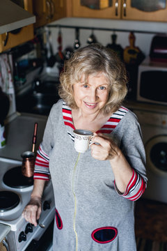 An elderly woman in home clothes holding mug of coffee in the kitchen