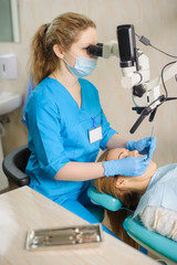 Naklejka na ściany i meble Young female dentist treating caries using microscope at the dentist office. Young woman patient lying on dentist chair with open mouth. Dentist wearing mask and gloves.