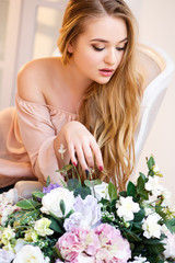 Young blonde woman surrounded with flowers in a light room. Tender portrait in a morning