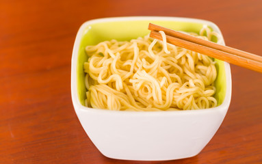 Elegant presentation of cooked noodles inside small square bowl sitting on wooden surface, sticks grabbing food, blurry background