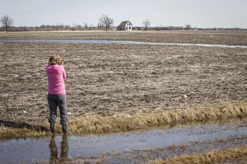 horizontal image of a woman wearing a pink sweater and grey slacks is standing in a field in water taking a picture of an old house in the distance in the spring time.