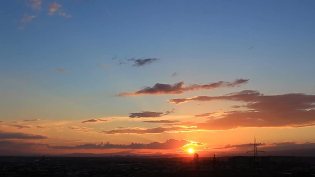 Sunset time-lapse with orange sun, blue sky, orange, yellow, grey clouds in the silhouette of the town