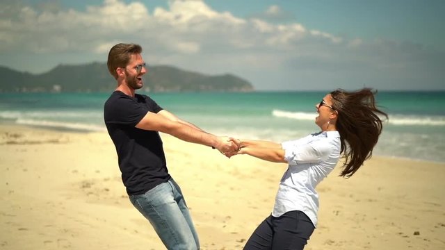 happy young couple holding hands and turning in slow motion on sandy beach in spring, buildings of seaside resort in background
