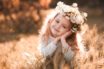 Smiling baby girl 3-4 year old wearing flower wreath outdoors. Sitting in meadow. Looking at...