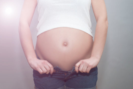 Pregnant girl in unbuttoned jeans and white t-shirt holding on to naked belly. The studio with gray background